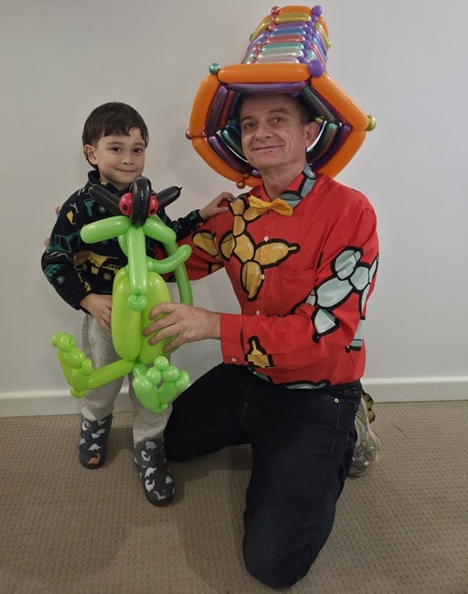 Adelaide Balloon Man wearing a colorful shirt and hat, posing alongside a young boy holding a green balloon dragon sculpture